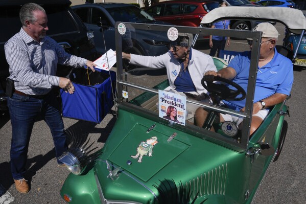 A couple drops their mail-in ballots into a collection box held by Sumter County Supervisor of Elections Bill Keen, during a golf cart parade to deliver mail-in votes organized by the Villages Democratic Club, in The Villages, Fla., Monday, Oct. 14, 2024. (AP Photo/Rebecca Blackwell)