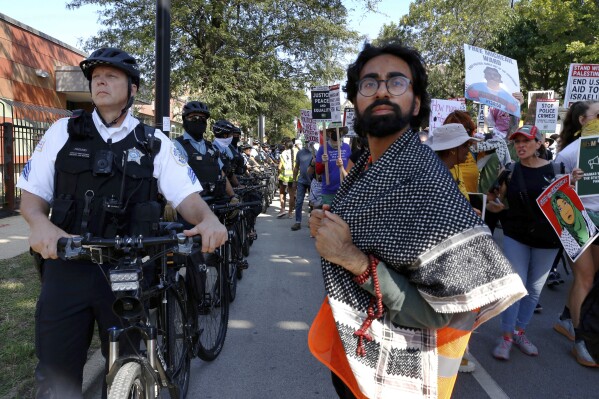 YM Masood, 20, right, stands next to a line of Chicago police officers during a March on DNC protest on Monday, Aug. 19, 2024, in Chicago, as he serves as a volunteer marshal for several of the protests to help keep protesters organized and safe and deescalate potential conflict. (AP Photo/Martha Irvine)