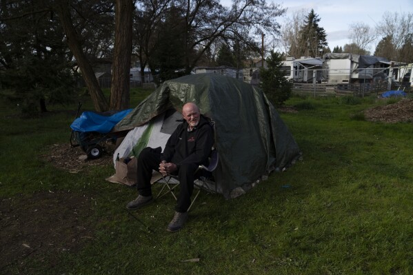 FILE - David Wilson sits outside his tent at Riverside Park on Thursday, March 21, 2024, in Grants Pass, Ore. The small Oregon city at the heart of a recent U.S. Supreme Court ruling on outdoor sleeping bans has voted to prohibit camping but establish certain areas where homeless people can go. (AP Photo/Jenny Kane, File)