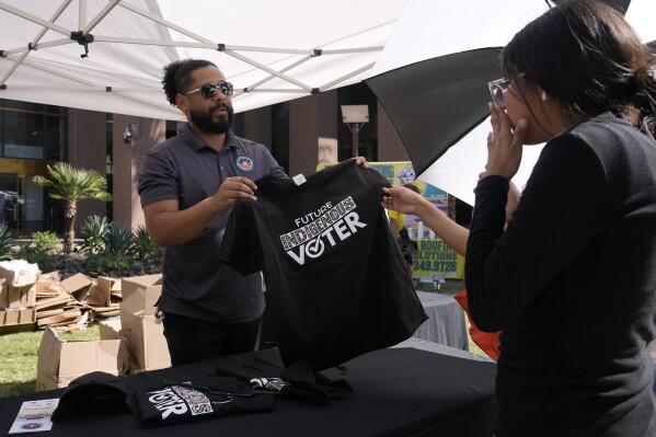 Alexander Castillo-Nunez, left, a civic engagement coordinator at the Inter Tribal Council of Arizona, Inc., shows a voter t-shirt at an Arizona Native Vote booth during an Indigenous Peoples' Day event Monday, Oct. 14, 2024, in Phoenix. (AP Photo/Ross D. Franklin)