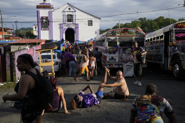 El peregrino Ernesto Troya estira la pierna de una amiga mientras descansan al dirigirse de rodillas hacia la iglesia de San Felipe, al fondo, para honrar al Cristo Negro en Portobelo, Panamá, el lunes 21 de octubre de 2024, durante un festival que celebra la icónica estatua que fue encontrada en la orilla del otrora puerto colonial en 1658. (Foto AP/Matías Delacroix)