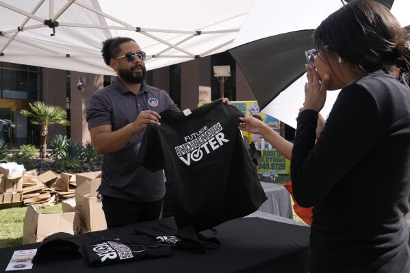 Alexander Castillo-Nunez, izquierda, coordinador de participación cívica en el Consejo Intertribal de Arizona, Inc, muestra una camiseta de votante en un puesto de Arizona Native Vote durante un evento del Día de los Pueblos Indígenas el lunes 14 de octubre de 2024, en Phoenix. (AP Foto/Ross D. Franklin)